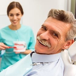 An older gentleman smiling while seated in the dentist’s chair and a hygienist shows him how to properly care for his dentures