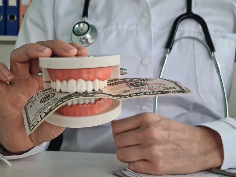 A jaw mockup biting a hundred-dollar bill, held in the hand of a dentist